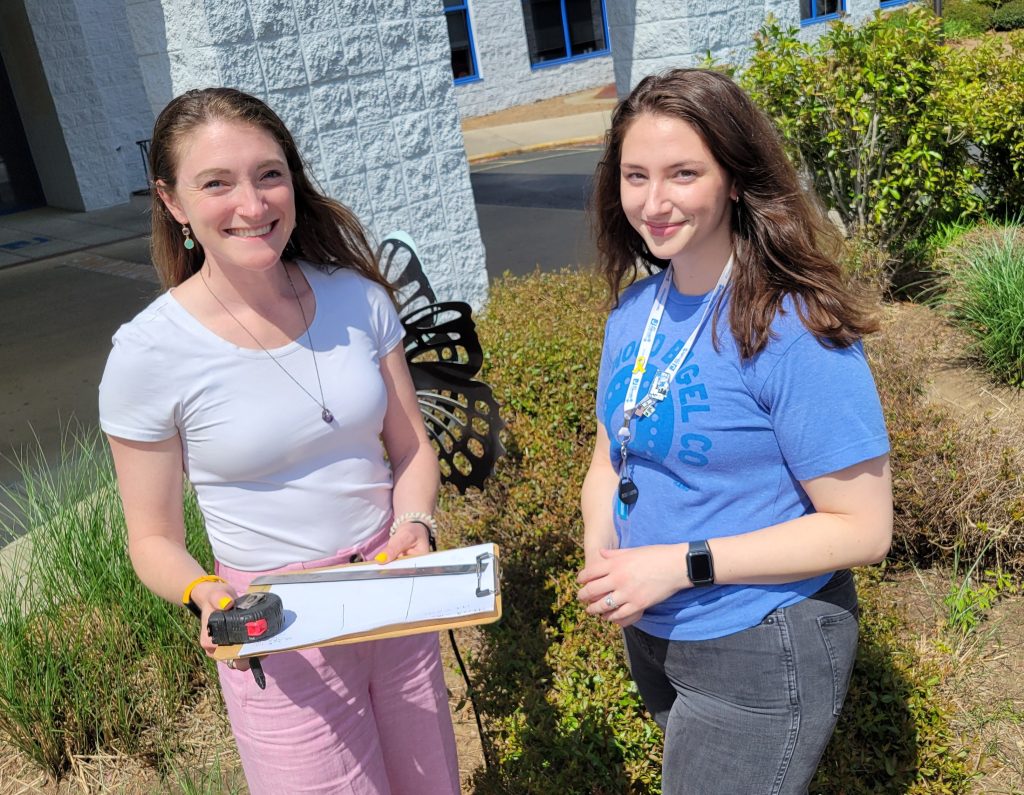Two women stand in a garden while consulting a clipboard