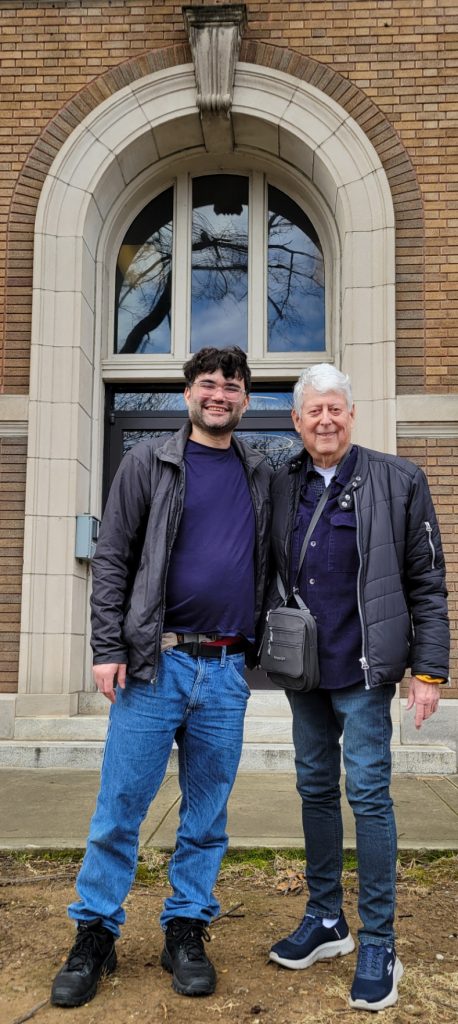 Two men pose in front of an orange brick building