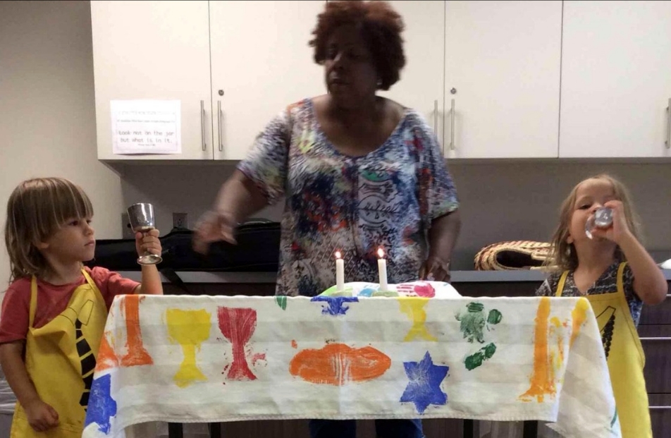 A woman stands behind a decorated table while one child on each end drinks from a chalice