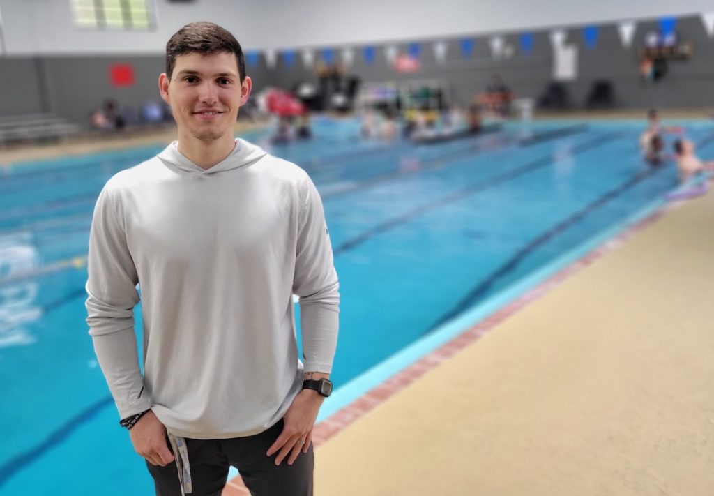 A man in a light gray top poses in front of The J's indoor pool while kids have swimming lessons in the background
