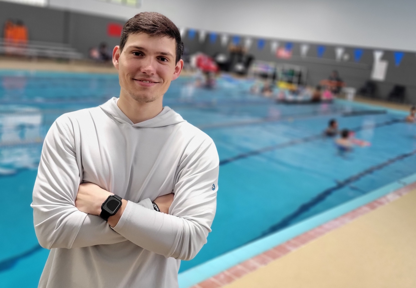 A man in a light gray top poses in front of The J's indoor pool while kids have swimming lessons in the background