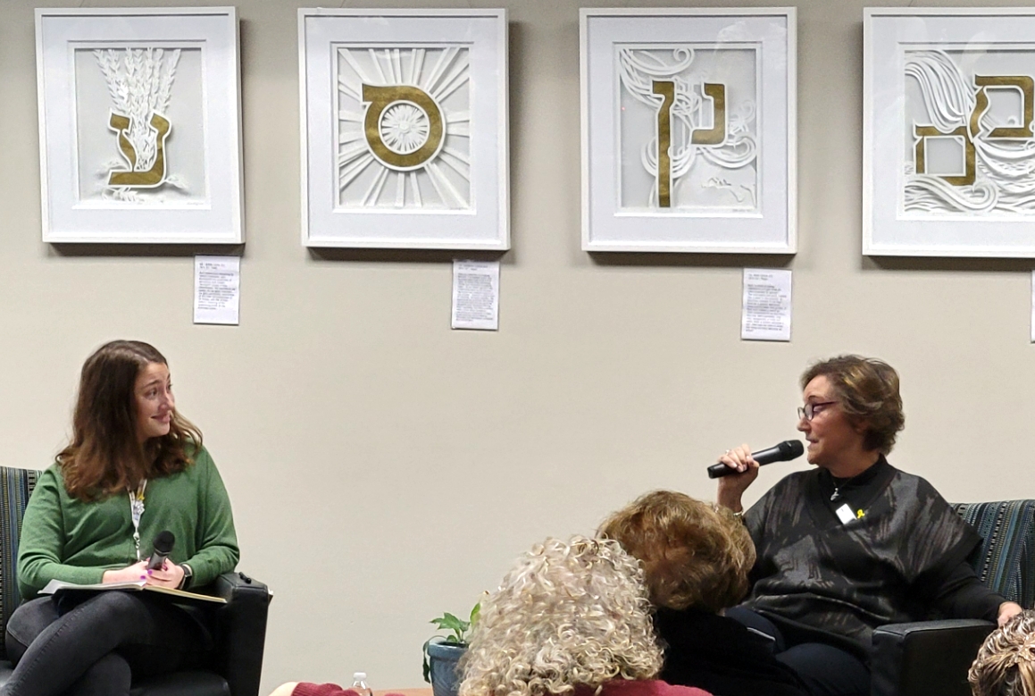 Two seated women face each other for an interview in front of a wall that's displaying artistic Hebrew letters