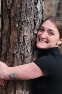 A woman smiles while hugging a tree in a forest
