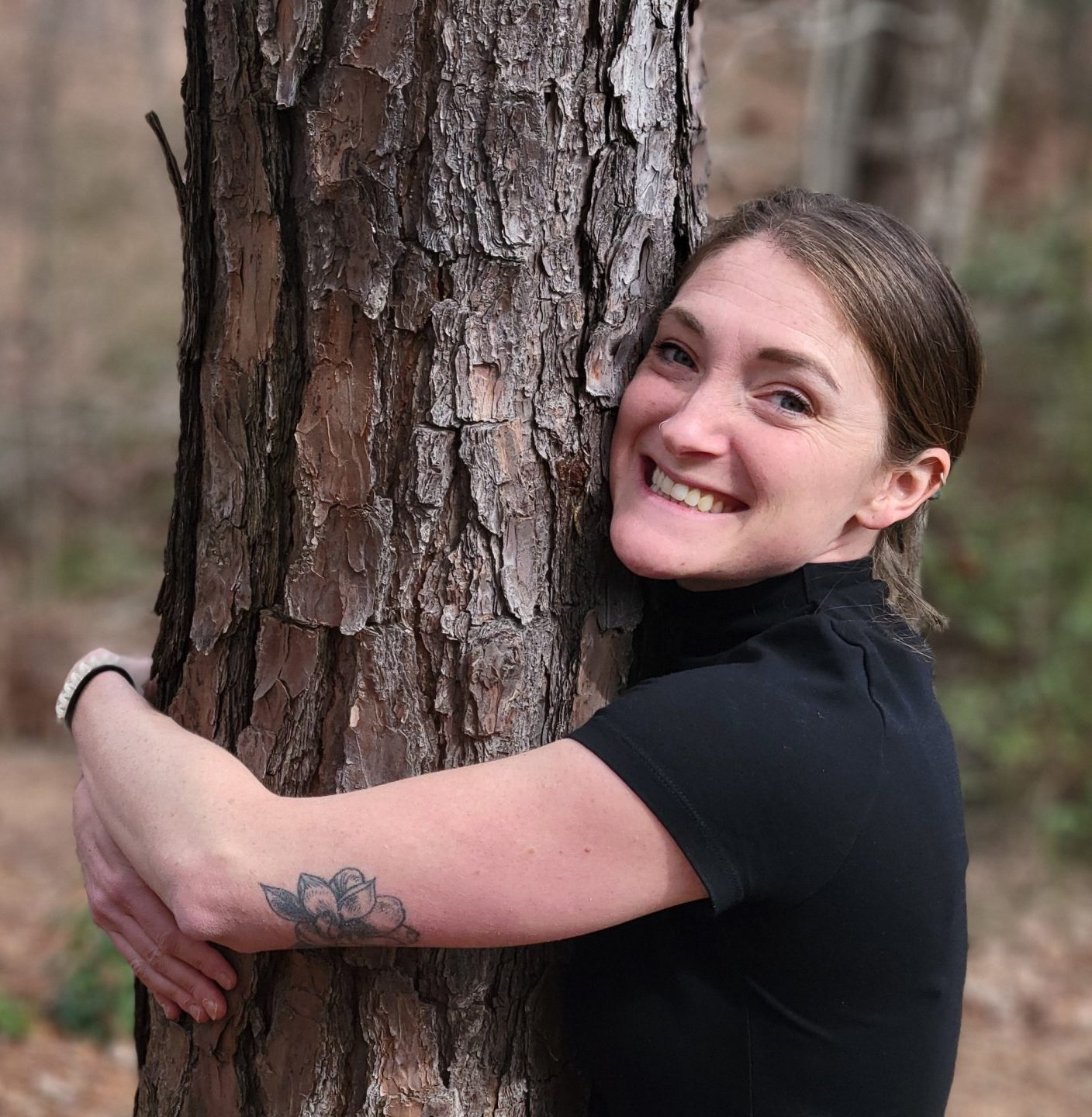 A woman smiles while hugging a tree in a forest