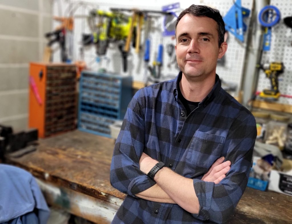 A man in blue checkered flannel crosses his arms as he leans back against a wooden work bench with a wall of tools behind him