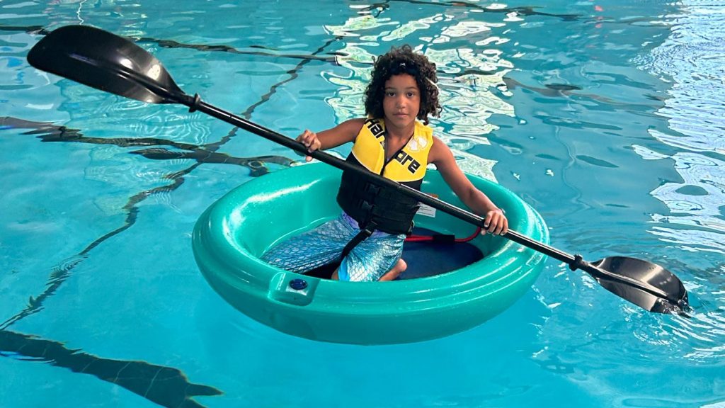 A young uses kayak paddles to propel a round boat in the indoor pool