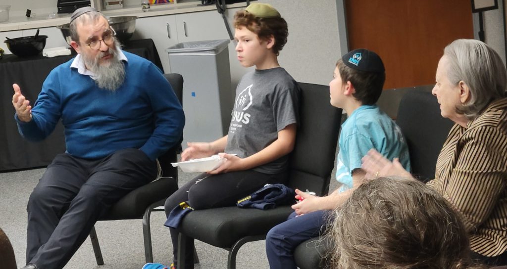 A rabbi, two children, and a woman are part of a larger group seated in a circle discussing Judaism and faith