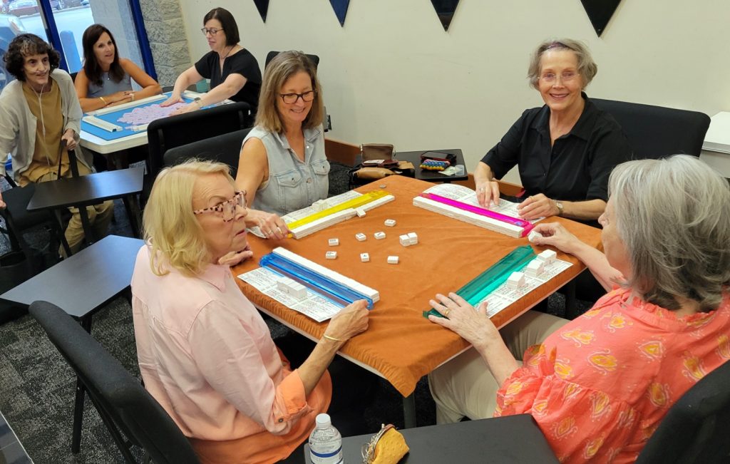 Four women in the foreground play mahjong at a table while another group plays in the background