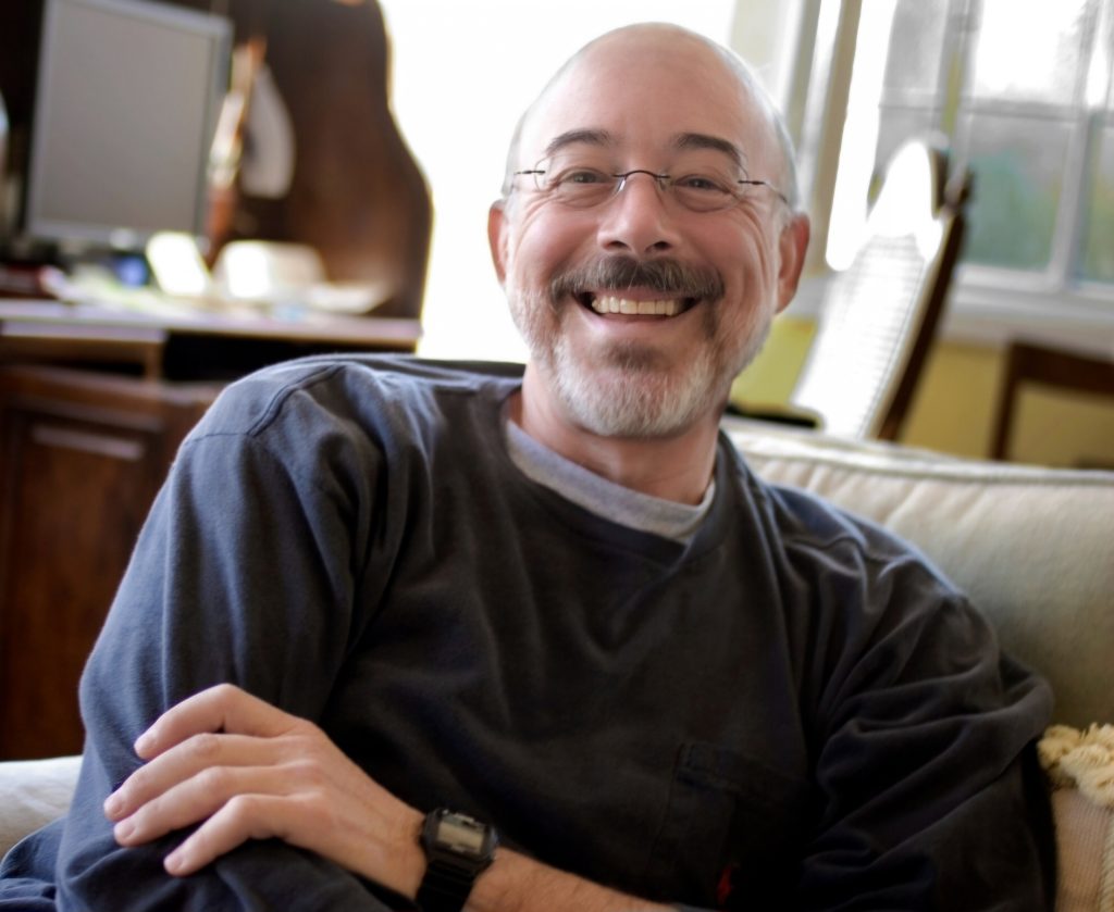 An affable bald man smiles and relaxes in his living room while looking at the camera