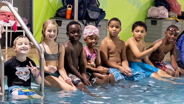 Young swimmers smile as they line up seated for a photo along the edge of The J's indoor pool