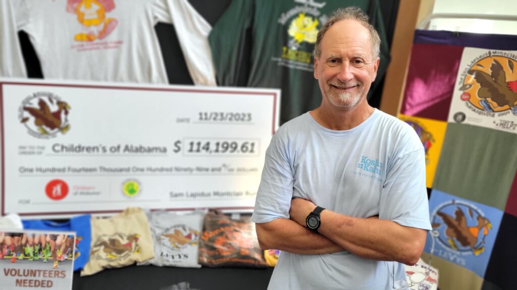 A man in a t-shirt poses in front of a display featuring a large check and shirts from past Sam Lapidus Montclair Run races