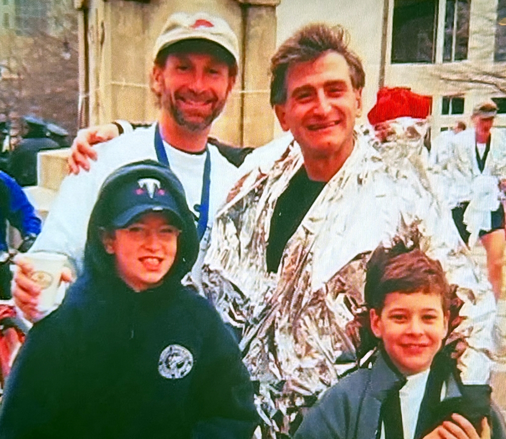 Two men (one wearing a cap and one in a space blanket) pose with two boys after a race