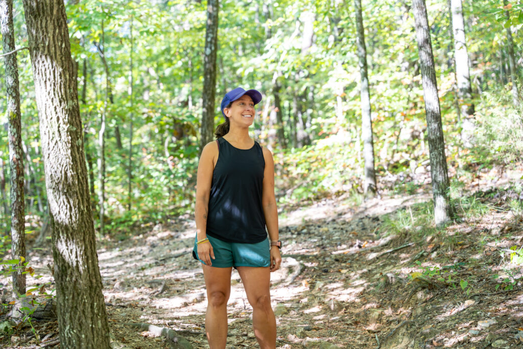 A woman in a ballcap, tank top, and shorts stands in the forest while admiring the LJCC's trail network.