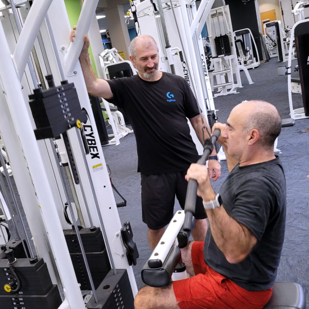 A member is seated at a fitness machine while his personal trainer looks on