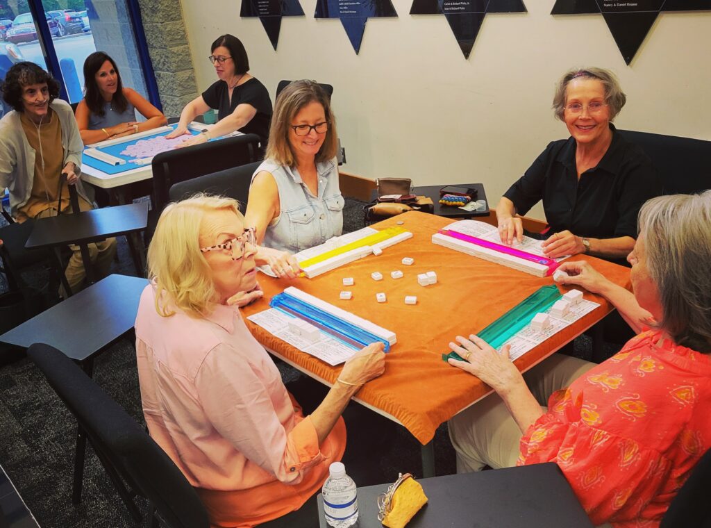 A group of seniors play mahjong at two tables in The J's lobby
