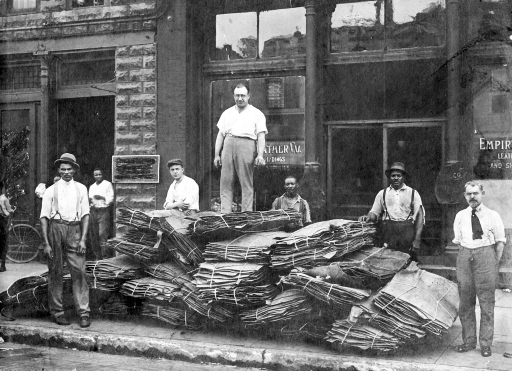 An old black and white photograph showing a group of men posing with a pile of bundled animal hides in front of a leather shop