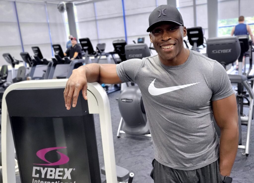A very fit man in a black cap poses by a piece of exercise equipment in a gym