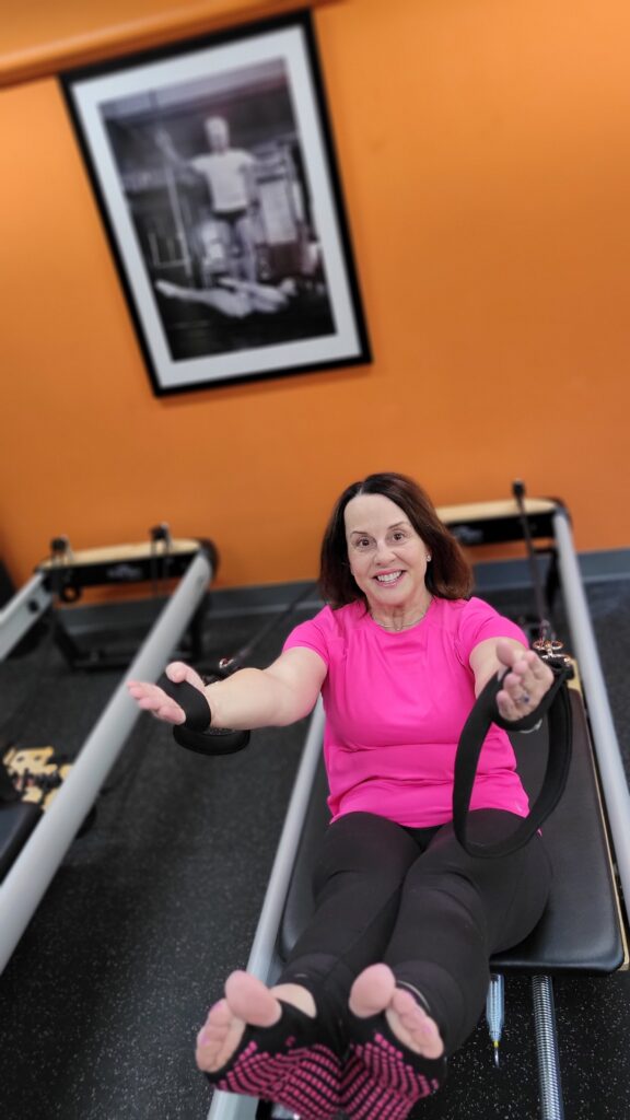 A woman in a pink top demonstrates one way to use a piece of Pilates reformer equipment that's on the floor