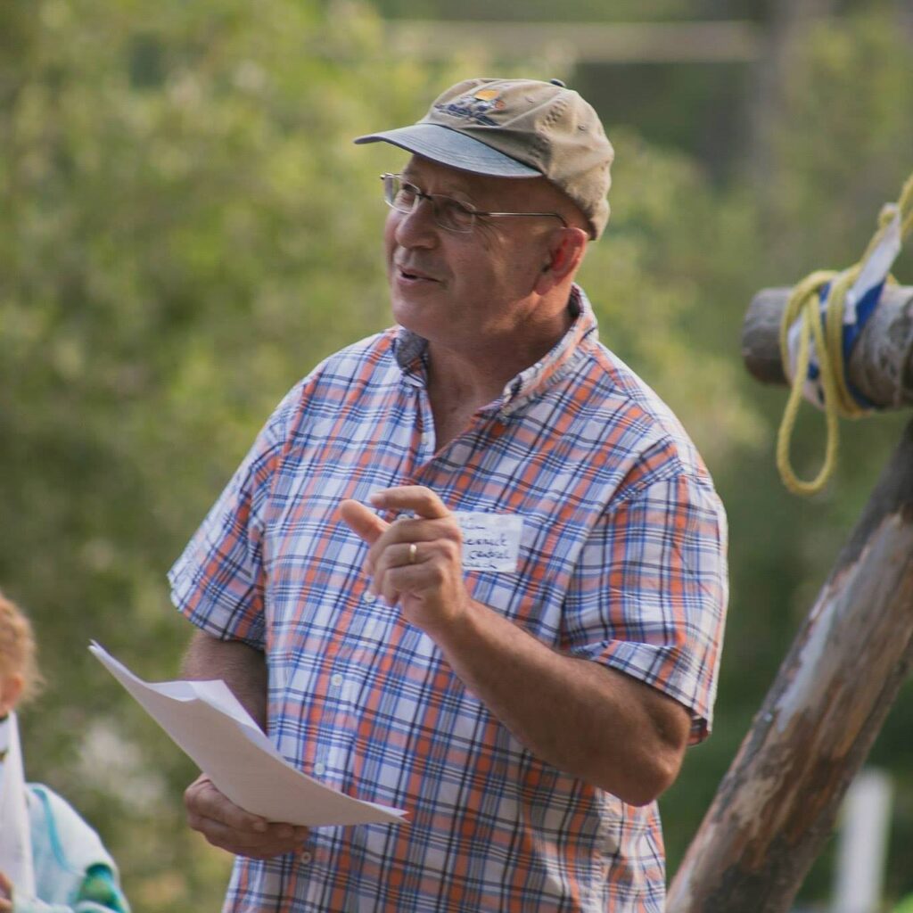 A man outside in a baseball-style cap and checkered shirt gestures with his thumb and forefinger