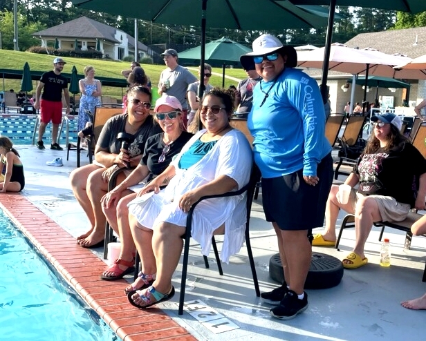 Three women are seated on chairs by a pool while a fourth stands next to them