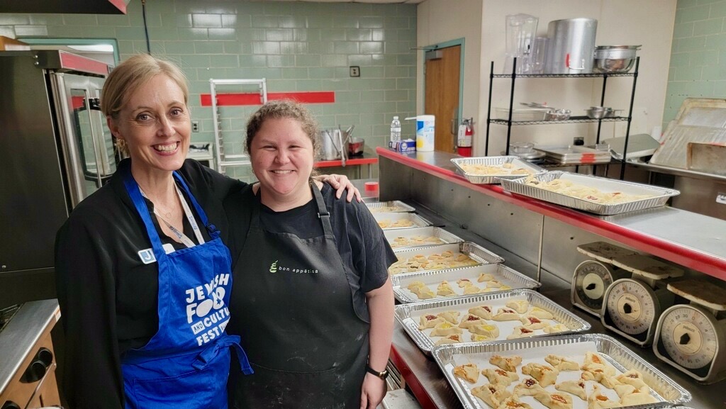 Two woman stand together in a kitchen with trays of freshly baked Hamantaschen cookies arrayed on the counter next to and behind them
