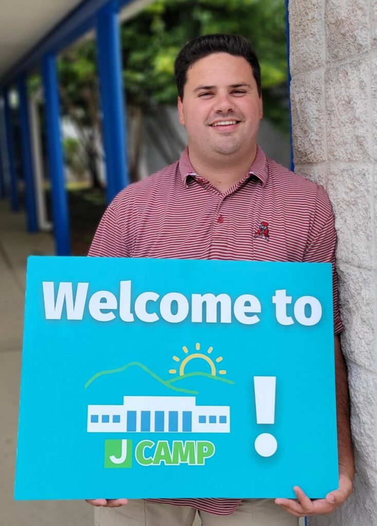 A man holds a "Welcome to J Camp" sign outside The J