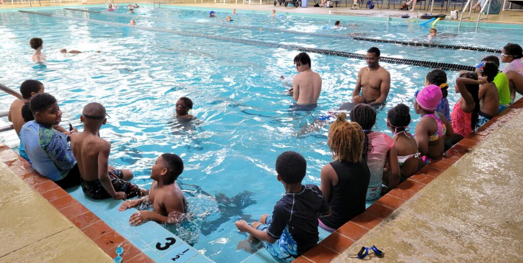 A group of young students sits on the edges of a corner of The J's indoor pool while swim instructors help other students in the water