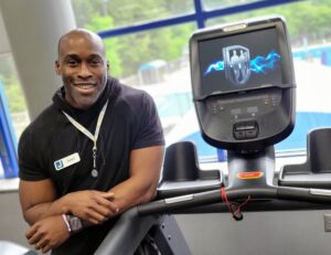 A fit bald man poses next to the display screen of a treadmill in front of a large window