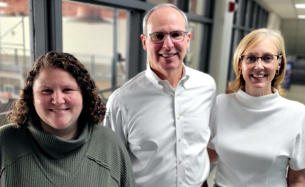 Two women flank a man as they all pose together in front of a wall of interior windows