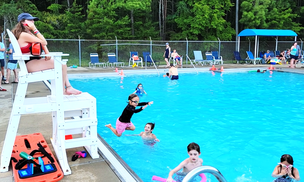 A lifeguard on a high white chair oversees children swimming in a large blue outdoor swimming pool