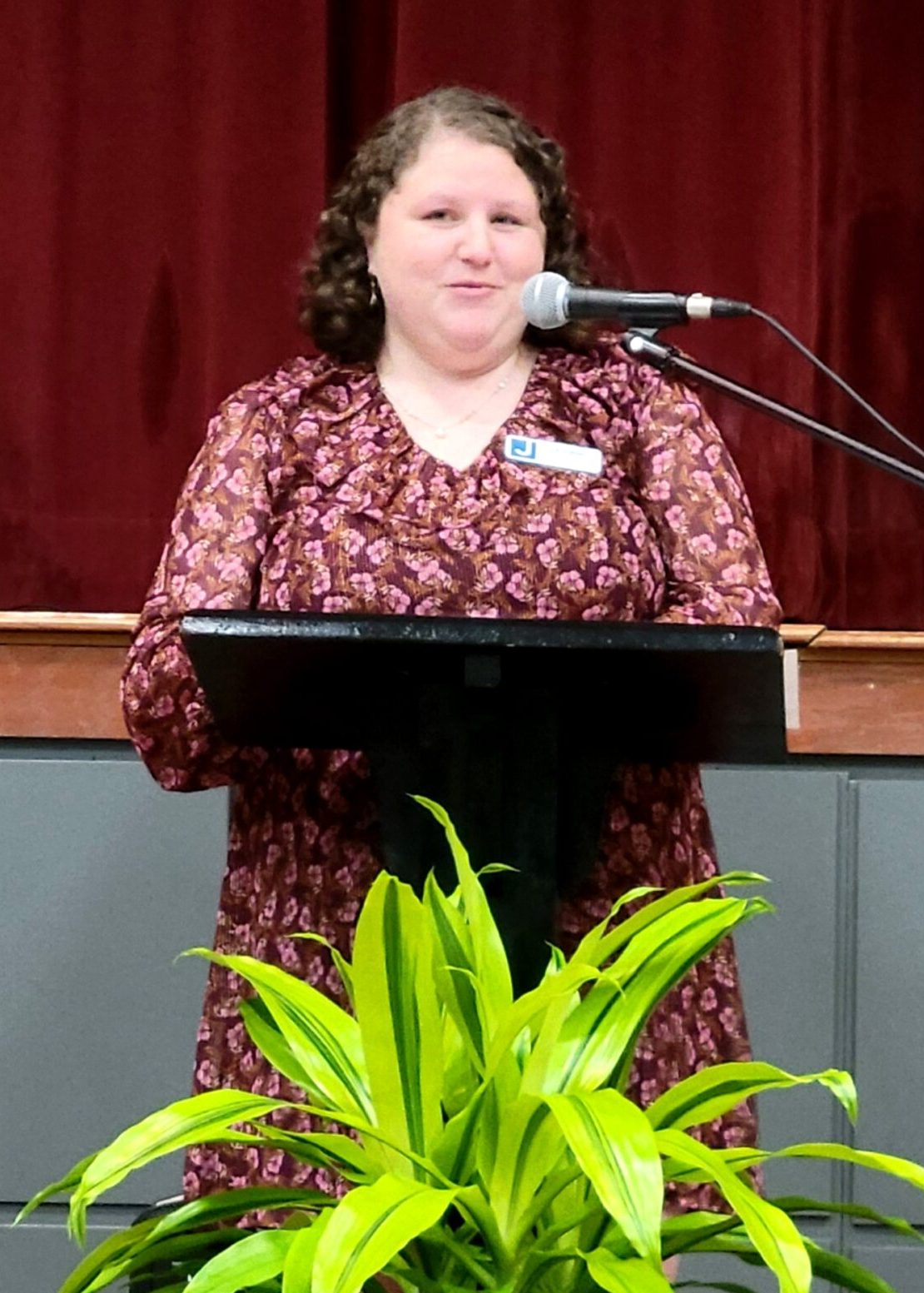 A woman in a dress stands at a podium that is flanked by some potted plants