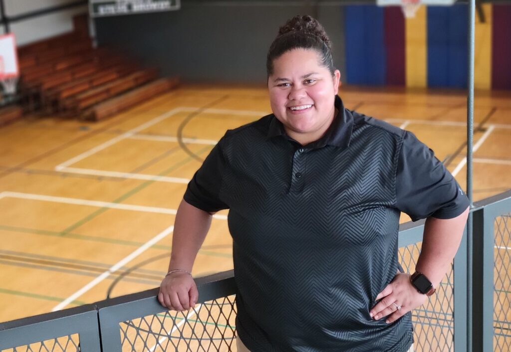A woman in a black polo shirt stands at a railing one floor above a gymnasium and bleachers that appear behind her.