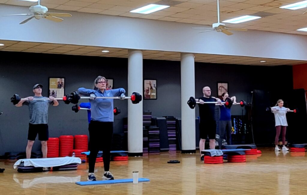 A group exercise class lifts weights in a large room with a wooden floor.