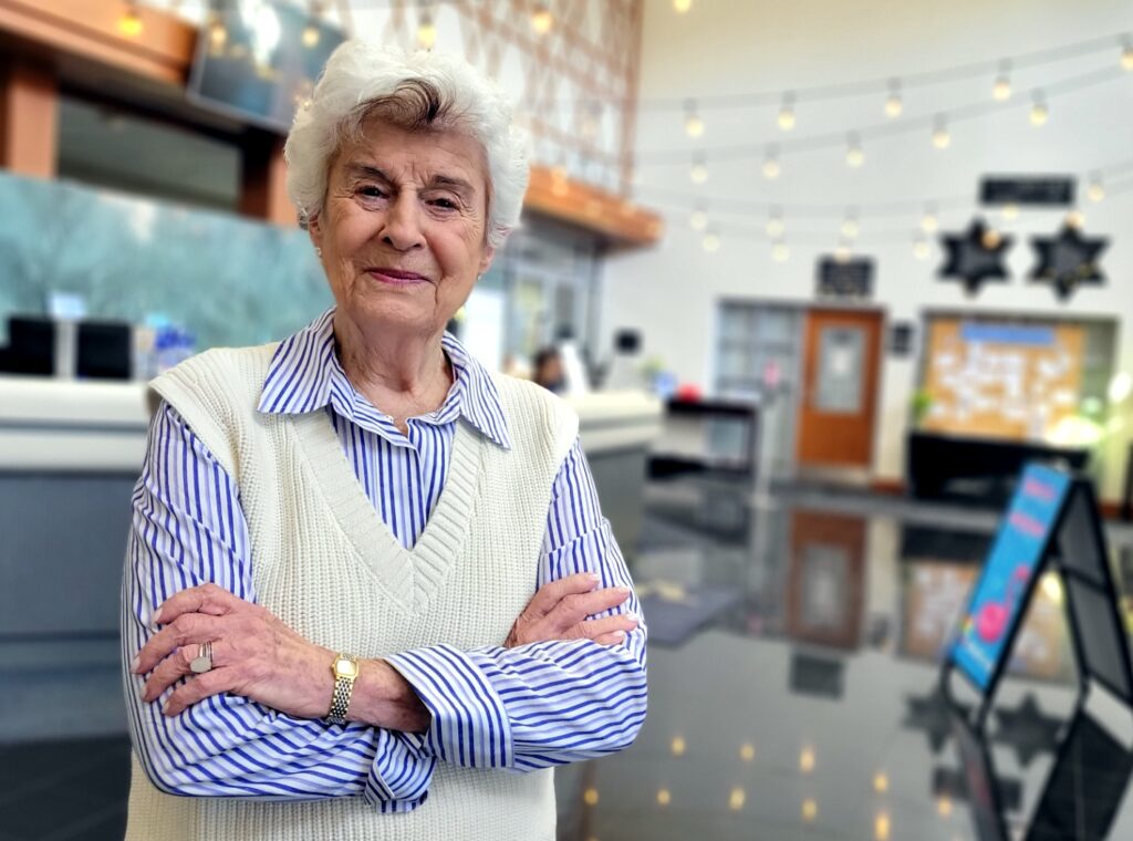 A woman with crossed arms, blue- and white-striped shirt, white vest and white hair poses in the lobby of The J