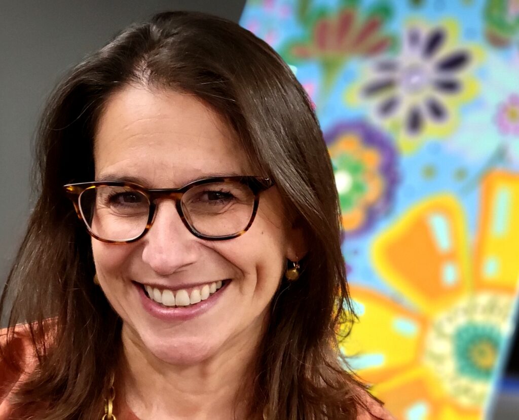 A woman with long hair and glasses poses in front of a painting of large individual flowers