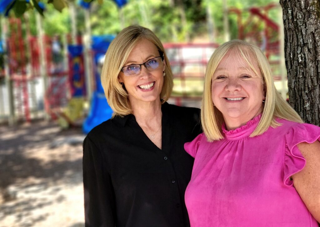 One woman in a black shirt and another woman in a fuchsia-colored dress stand next to each other against a tree, with playground equipment in the background