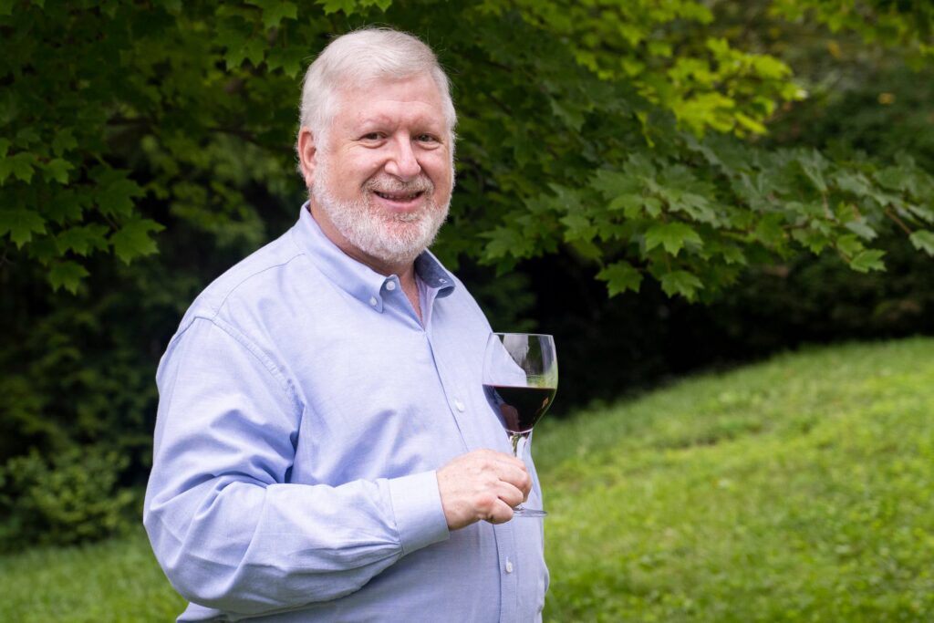 Bearded white-haired man in light-blue long-sleeved shirt holds a glass of red wine with greenery in the background