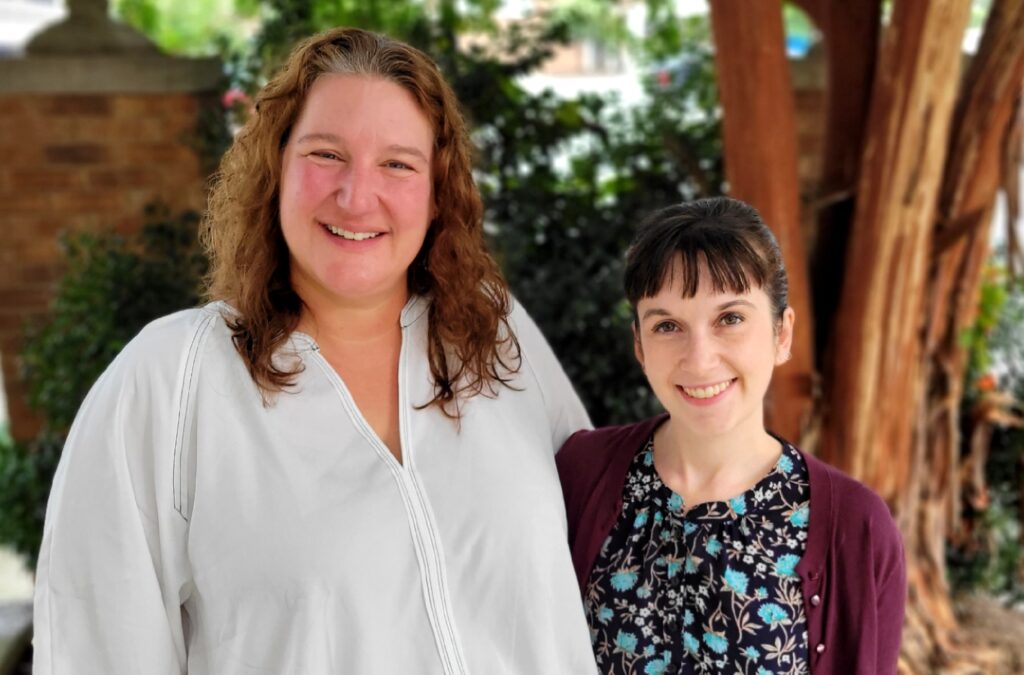 Two women (one taller in a white blouse and the other, at right, wearing a dark-patterned blouse) pose in front of a tree and some bushes