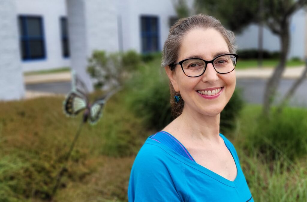A smmiling woman in a blue shirt and glasses stands in front of The J's butterfly garden