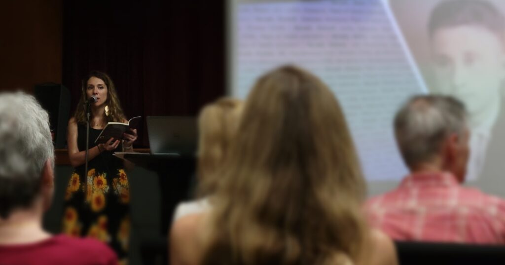 A woman holds a book at a podium while a screen behind her projects the image of her great grandfather. Audience members are seated in the foreground.