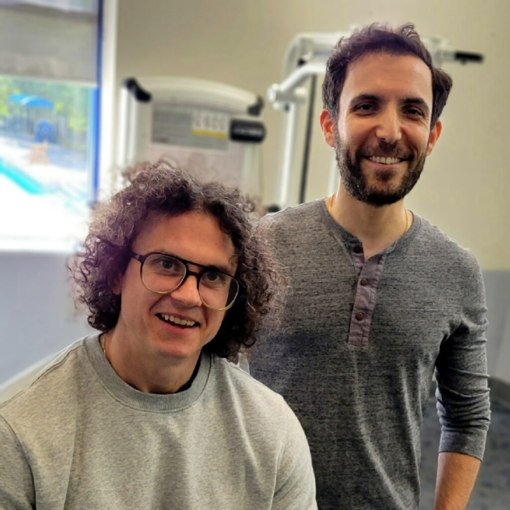 Two men, one seated with very curly hair and one bearded and standing at right, pose in front of some exercise equipment and a window.