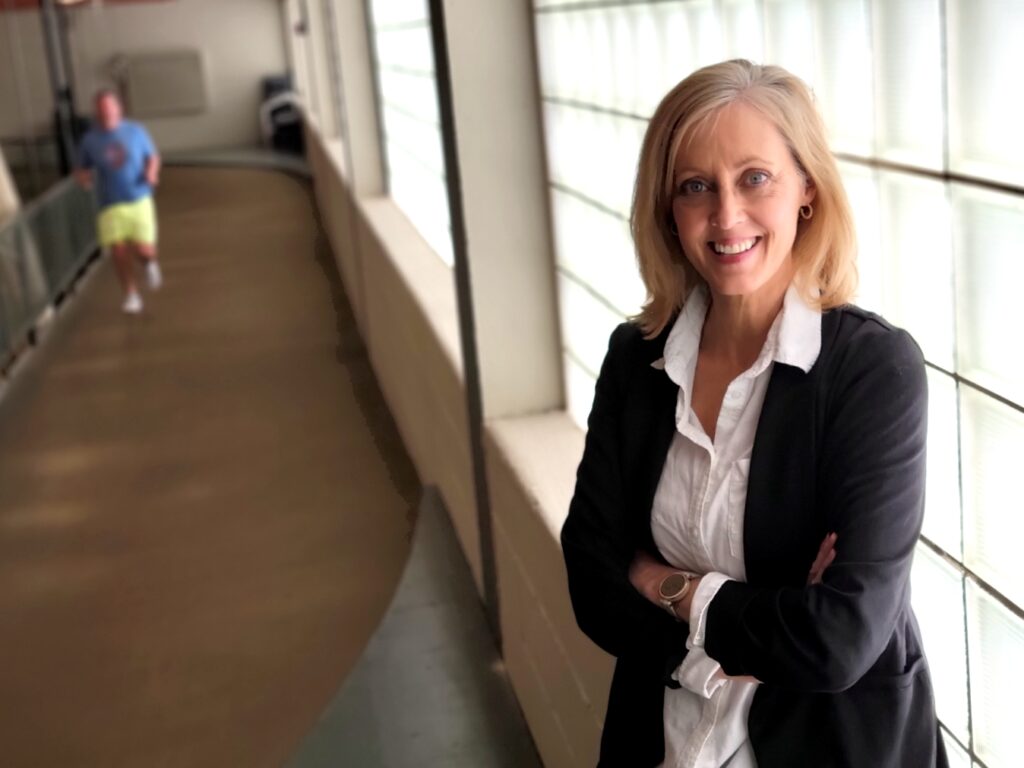 A woman in a white shirt and dark jacket poses in front of a glass-block wall by an indoor track. In the background, a man is running on the track.