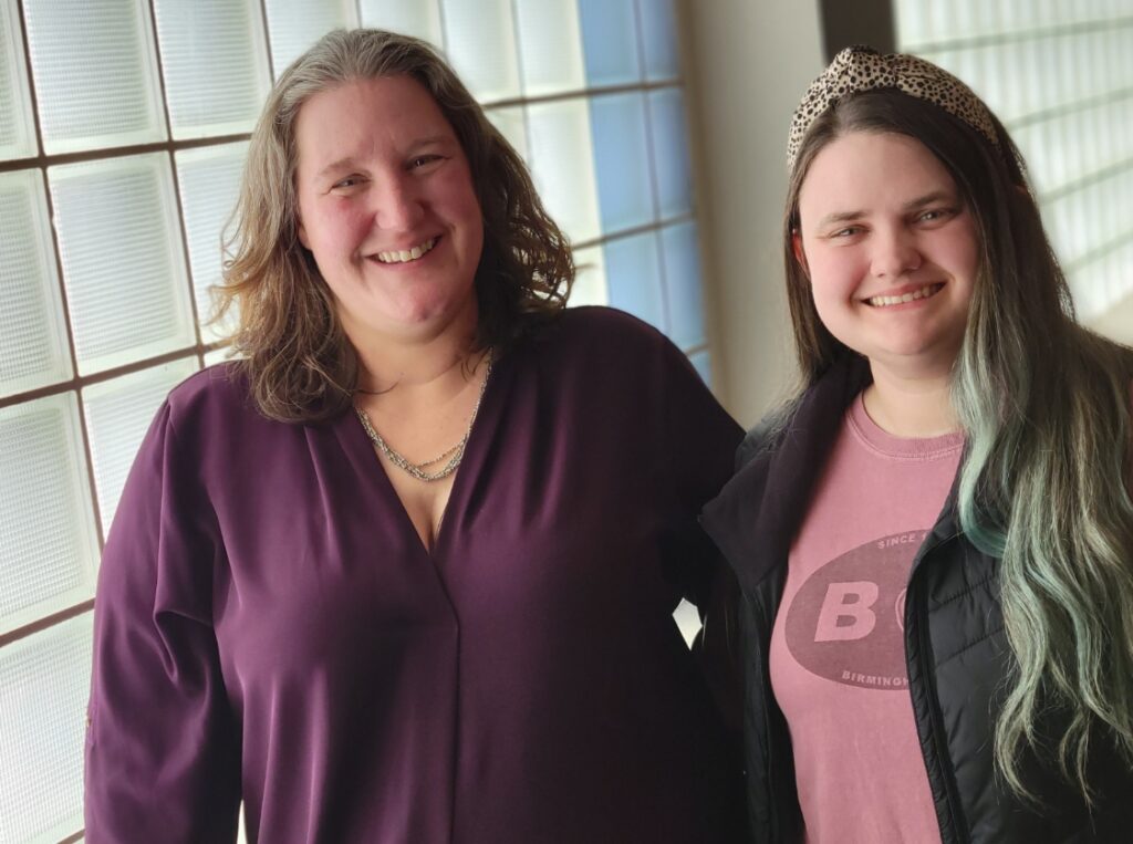 Two smiling women stand next to each other in front of a glass wall