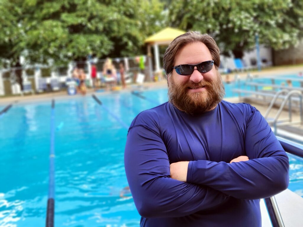 A man in sunglasses and long-sleeved shirtposes in front of an outdoor swimming pool
