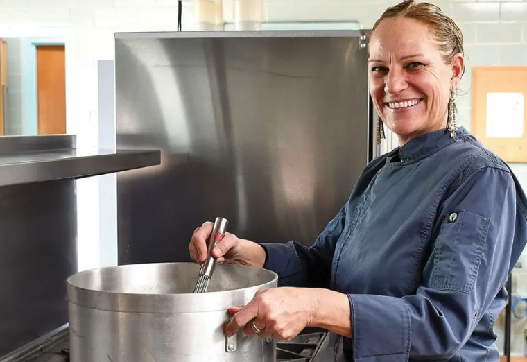 A woman in a gray chef's jacket poses by a large pot in The J's kosher kitchen
