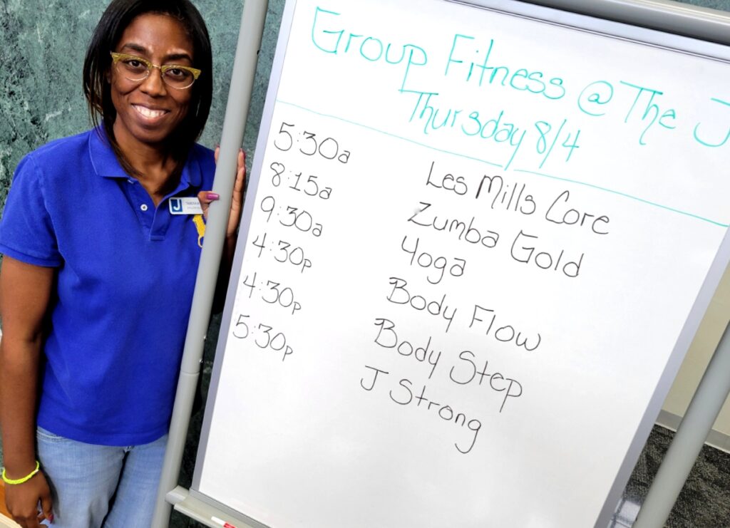 A woman stands next to the large white board that she used to write down the day's Group Fitness schedule