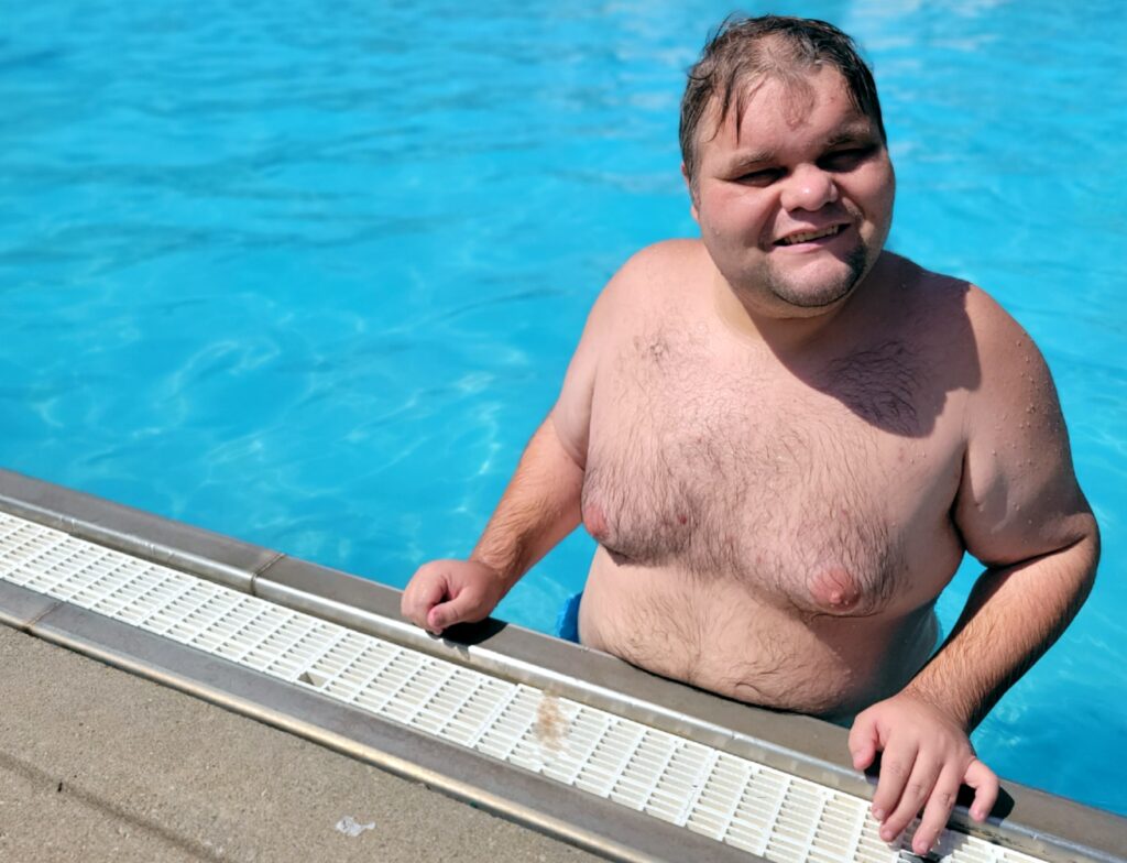 A man in a bathing suit smiles into the camera while standing in the water by the edge of a swimming pool