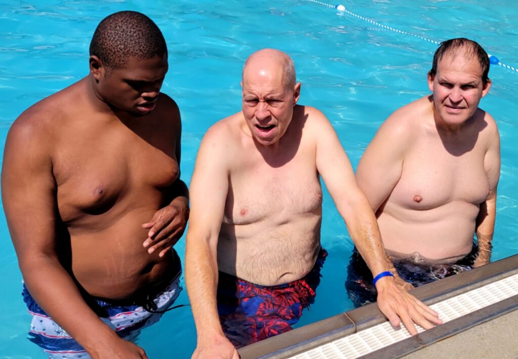 Three men in bathing suits pose in the water by the edge of a swimming pool