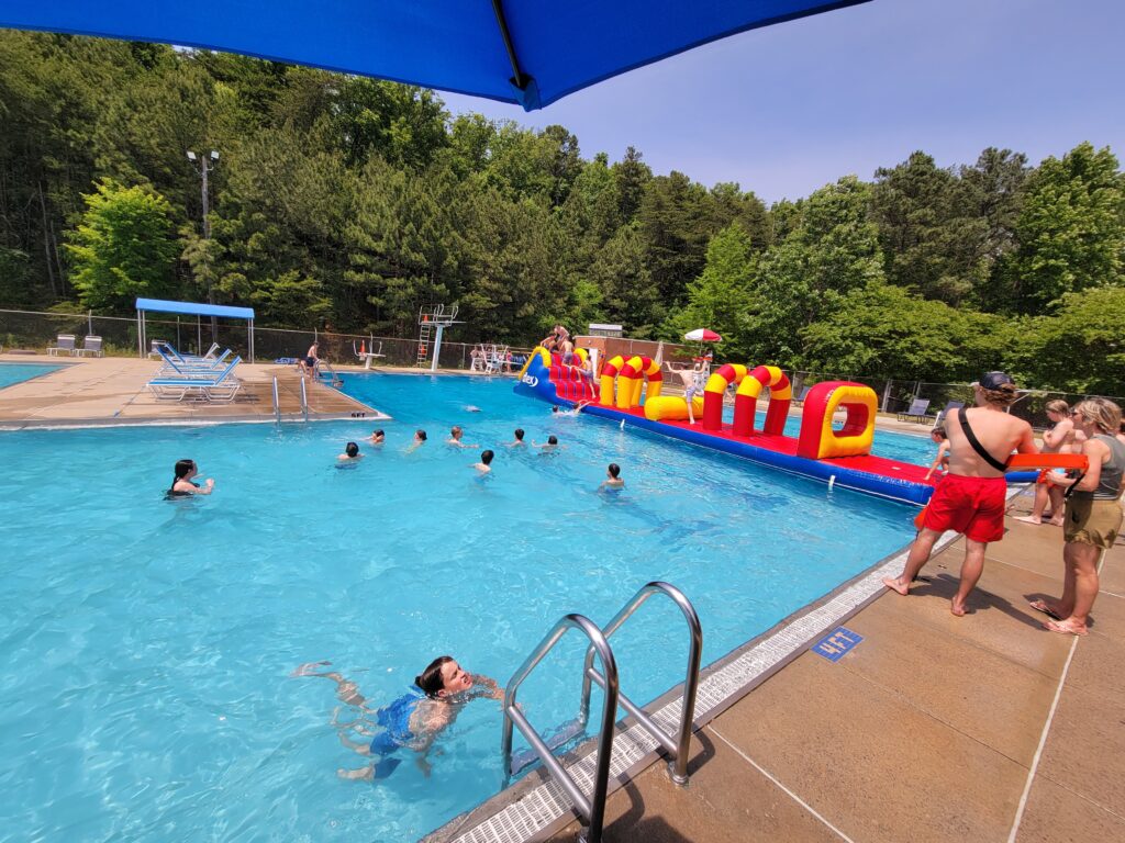 A picture of a large outdoor pool with kids swimming in the foreground and playing on a giant blue, red, and yellow inflatable in the background