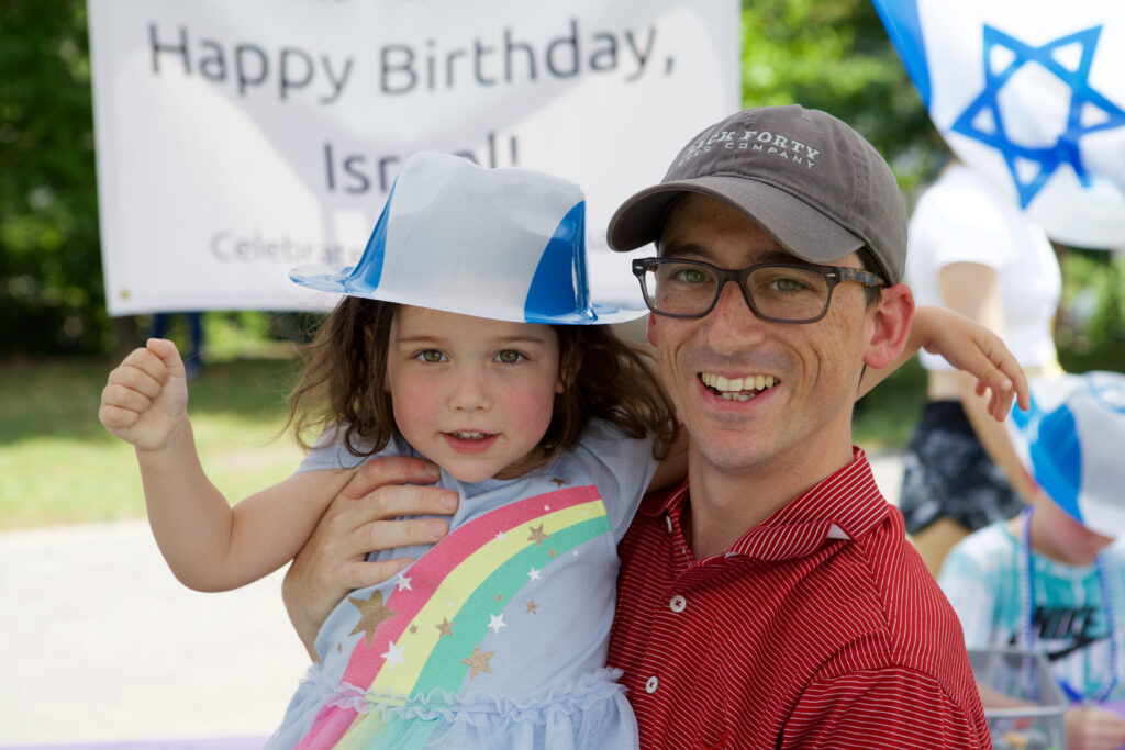 A man holds a young girl in a blue and white hat as they stand in front of a banner celebrating Israeli independence day, or Yom Ha'Atzmaut